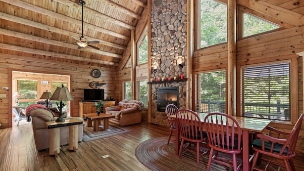 A wooden cabin interior with a stone fireplace, wooden furniture, and large windows. - Luxury Log Cabin • Paradise in the Lakes Region (Naples)