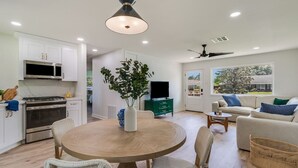 A modern kitchen with white cabinets, a built-in microwave, and a stove. There is a dining area with a round table and wicker chairs. A TV is on a green stand, and a sofa is in the living room with a view of the outdoors. - KING Bed/Central Location/Fast wi-fi/5star listing (Lafayette)