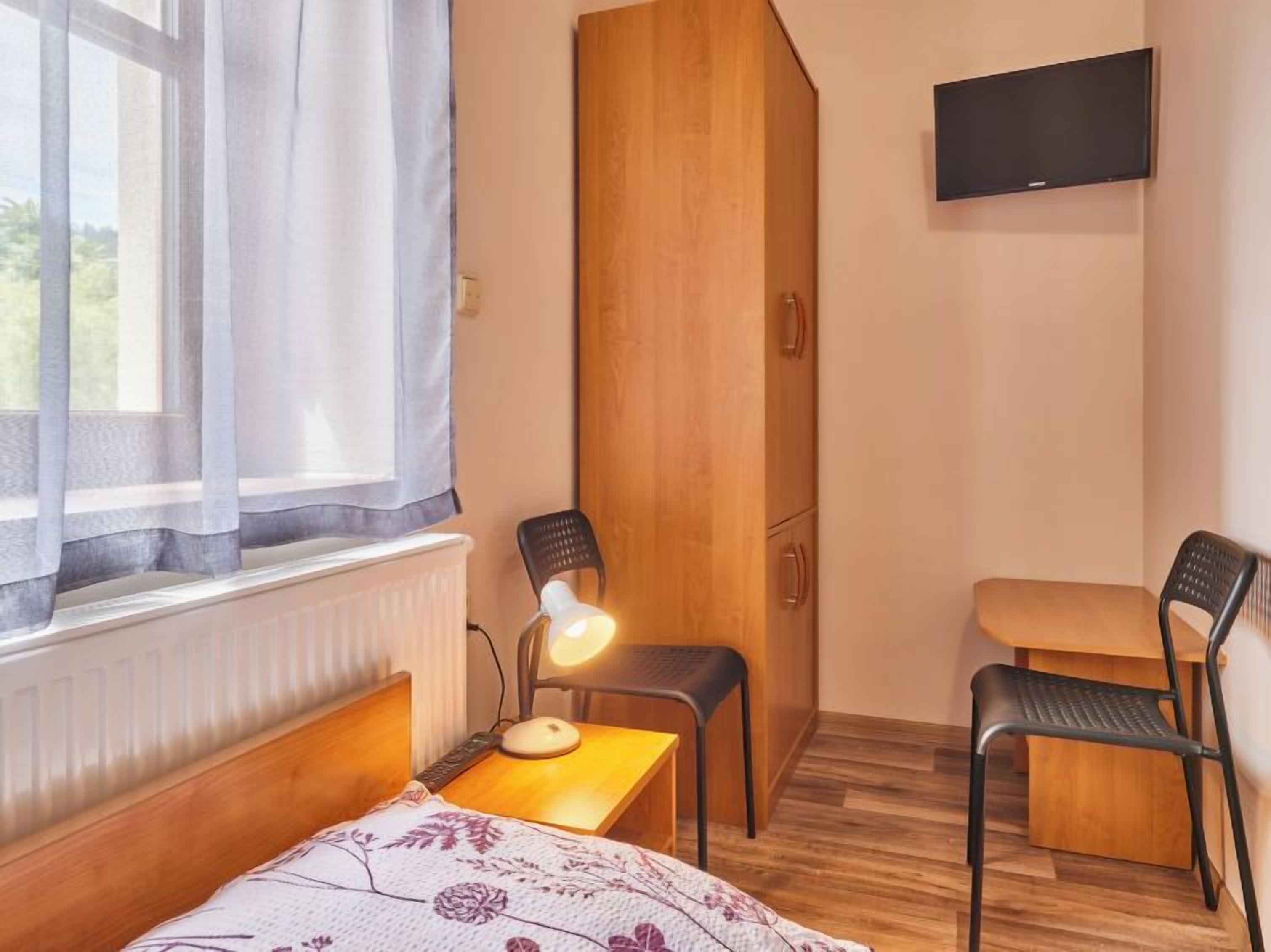 A compact room with a bed, a desk, two chairs, a wooden wardrobe, a radiator, a window with curtains, and a TV mounted on the wall.