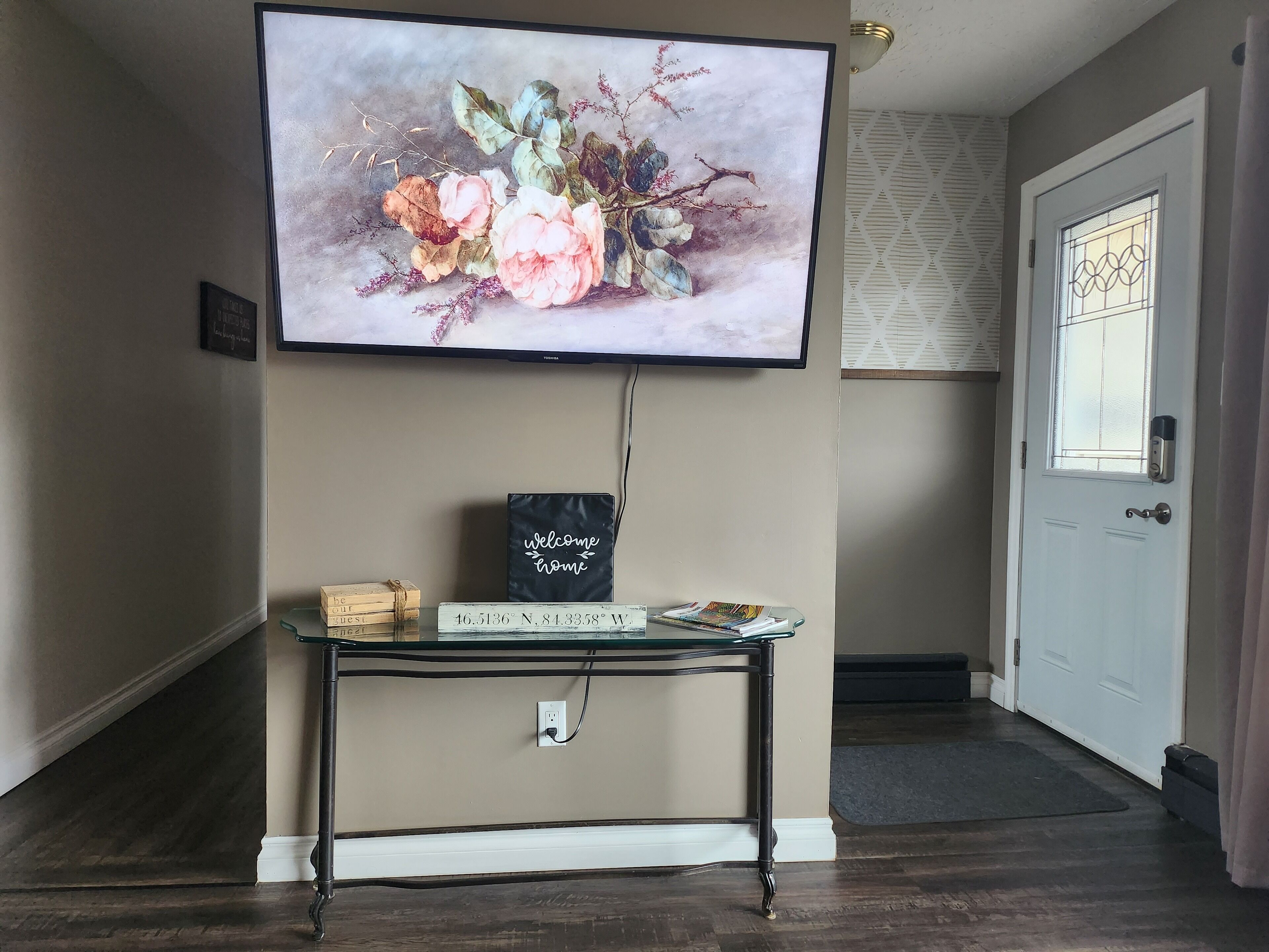 Una sala de estar con un televisor montado que muestra un cuadro floral, una mesita con un letrero de bienvenida y una puerta con un ventanal.