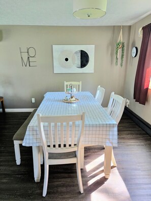 Un coin repas avec une table dressée pour quatre personnes, des chaises blanches, un banc, une horloge murale et une œuvre d’art décorative accrochée au mur.