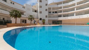 A swimming pool surrounded by a multi-story building with balconies, palm trees, and lounge chairs. - Apartment Delphine with pool (Porches)