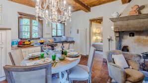 Ein Essbereich mit Holztisch, Stühlen und einer Chandelier. Im Hintergrund ist eine Küche mit Fenster und Heizkörper zu sehen.