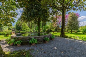 Ein runder Steinteich, umgeben von Kies, mit Kübelpflanzen und Bäumen in einer Gartenanlage.