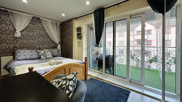 Een slaapkamer met een groot raam, een bed en uitzicht op een gebouw en groen.
