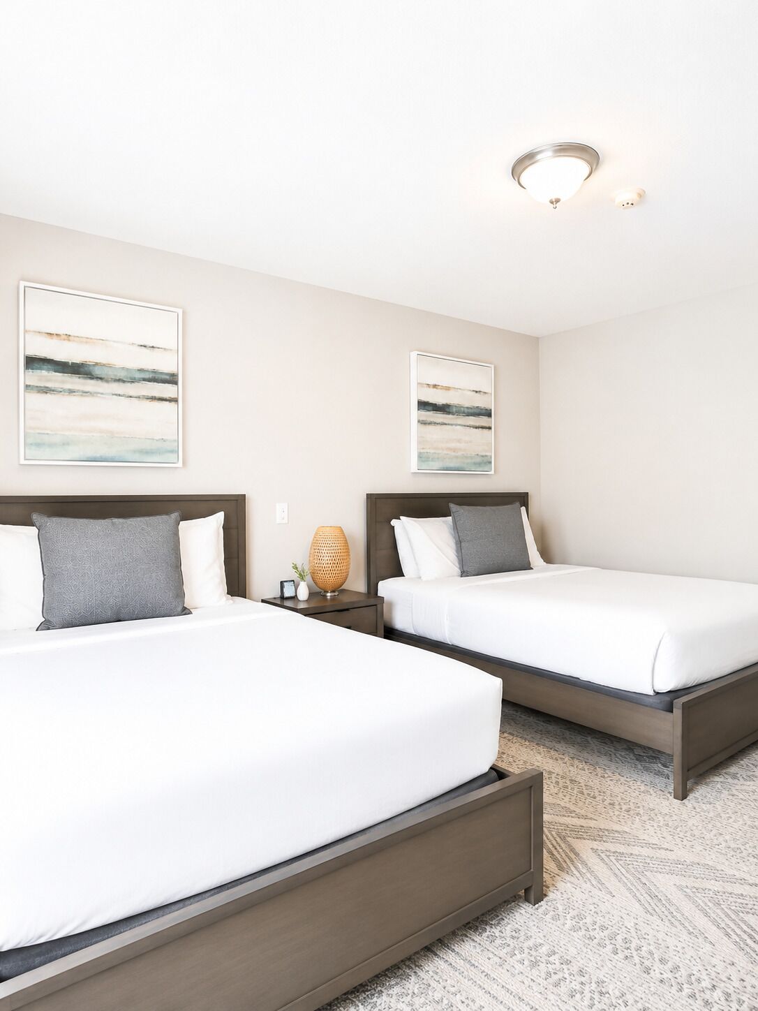 A hotel room with two beds, a nightstand, a lamp, and two framed abstract paintings on the wall.