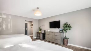 A hotel room with two beds, a desk with a chair, a dresser, a television mounted on the wall, and a potted plant.