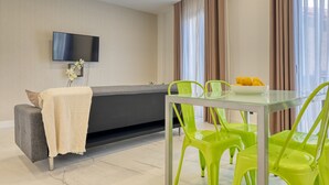 A modern living room with a sofa, a glass dining table, and bright green chairs. A flat-screen TV is mounted on the wall, and there is a small plant on the sofa.