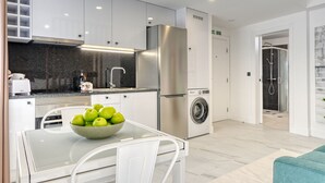 A modern kitchen with white cabinets, a stainless steel refrigerator, a washing machine, and a glass dining table with a bowl of green apples. - Beco Santa Emilia 4Q, a Home in Madeira (Funchal)