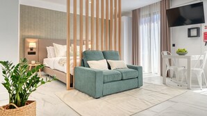 A modern hotel room with a green sofa, a bed with white linens, a small bedside table with a lamp, a dining area with a white table and chairs, and a large TV mounted on the wall. - Beco Santa Emilia 4Q, a Home in Madeira (Funchal)
