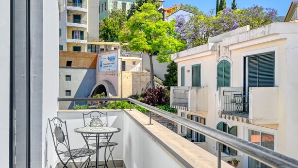A balcony with a table and chairs, overlooking a cityscape with buildings and a tree. - Beco Santa Emilia 4Q, a Home in Madeira (Funchal)