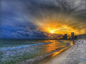 Ein Strand mit Wellen, einem Sonnenuntergang und einer Stadtansicht im Hintergrund.