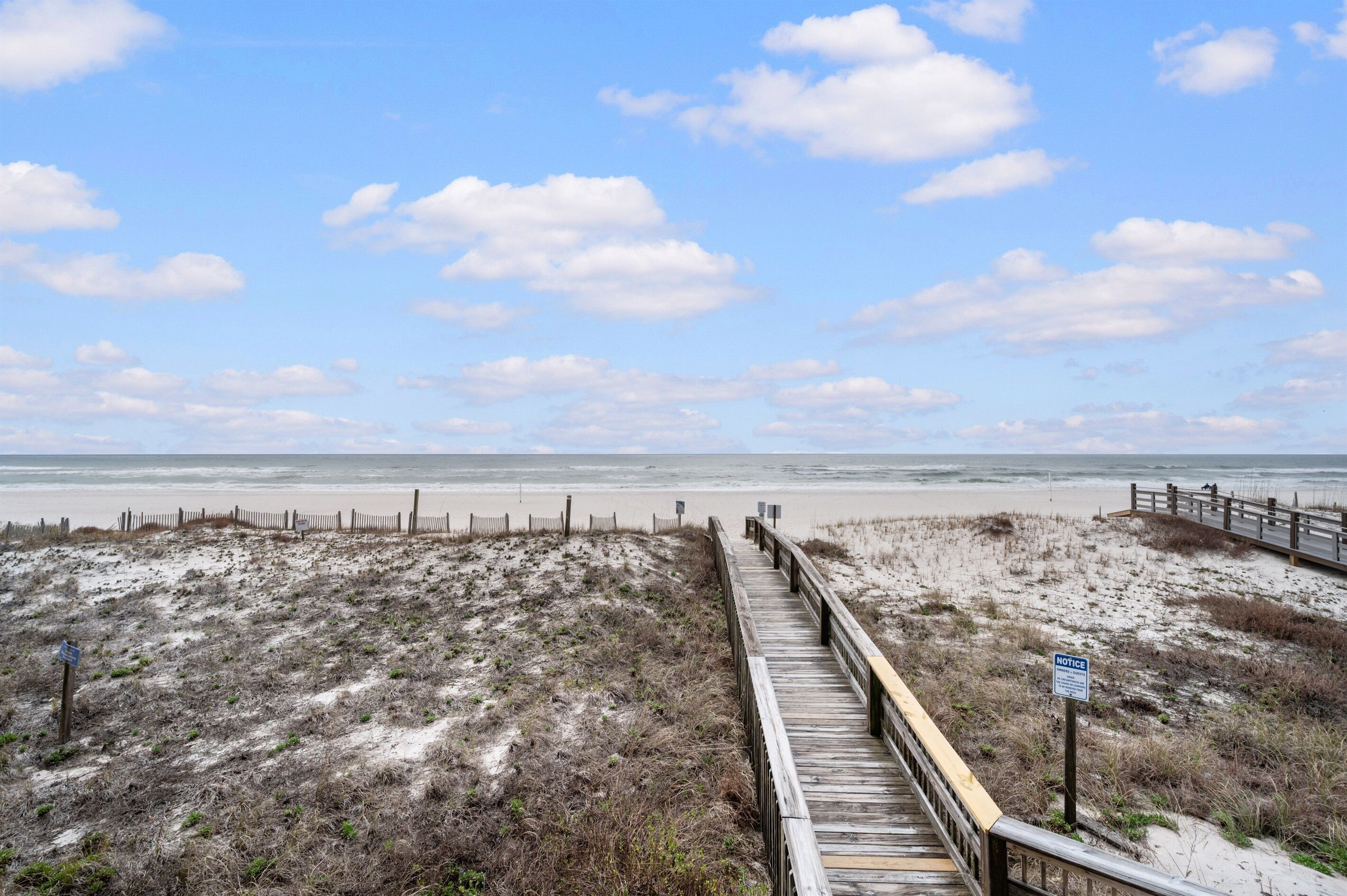 Une promenade en bois qui mène à une plage au paysage sablonneux, sous un ciel dégagé.