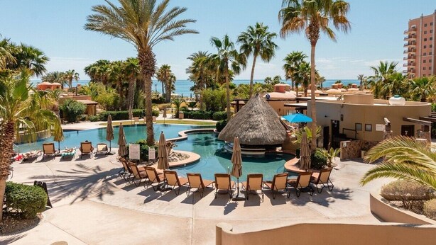 Zwembad van een resort met ligbedden en parasols, omringd door palmbomen en uitzicht op de oceaan.