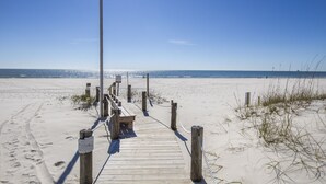Une promenade en bois mène à une plage de sable fin, sous un ciel bleu et dégagé.