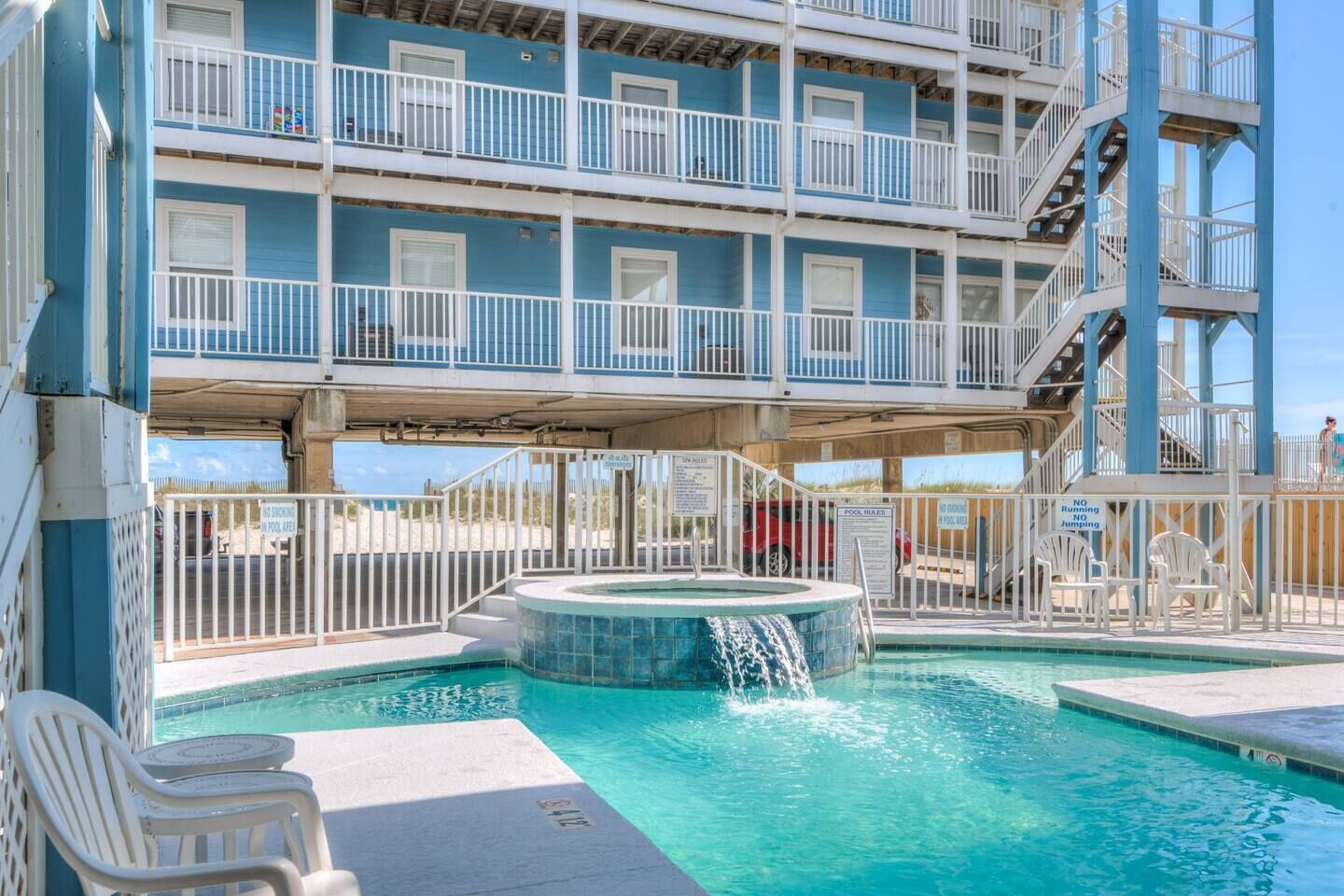 A swimming pool with a hot tub, surrounded by white chairs and a multi-story building with balconies.