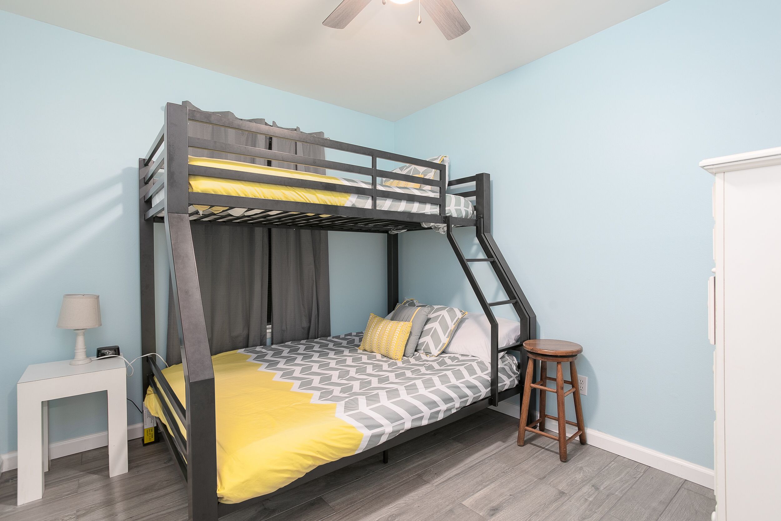 A bunk bed with a geometric-patterned bedspread, a small white nightstand with a lamp, and a wooden stool.