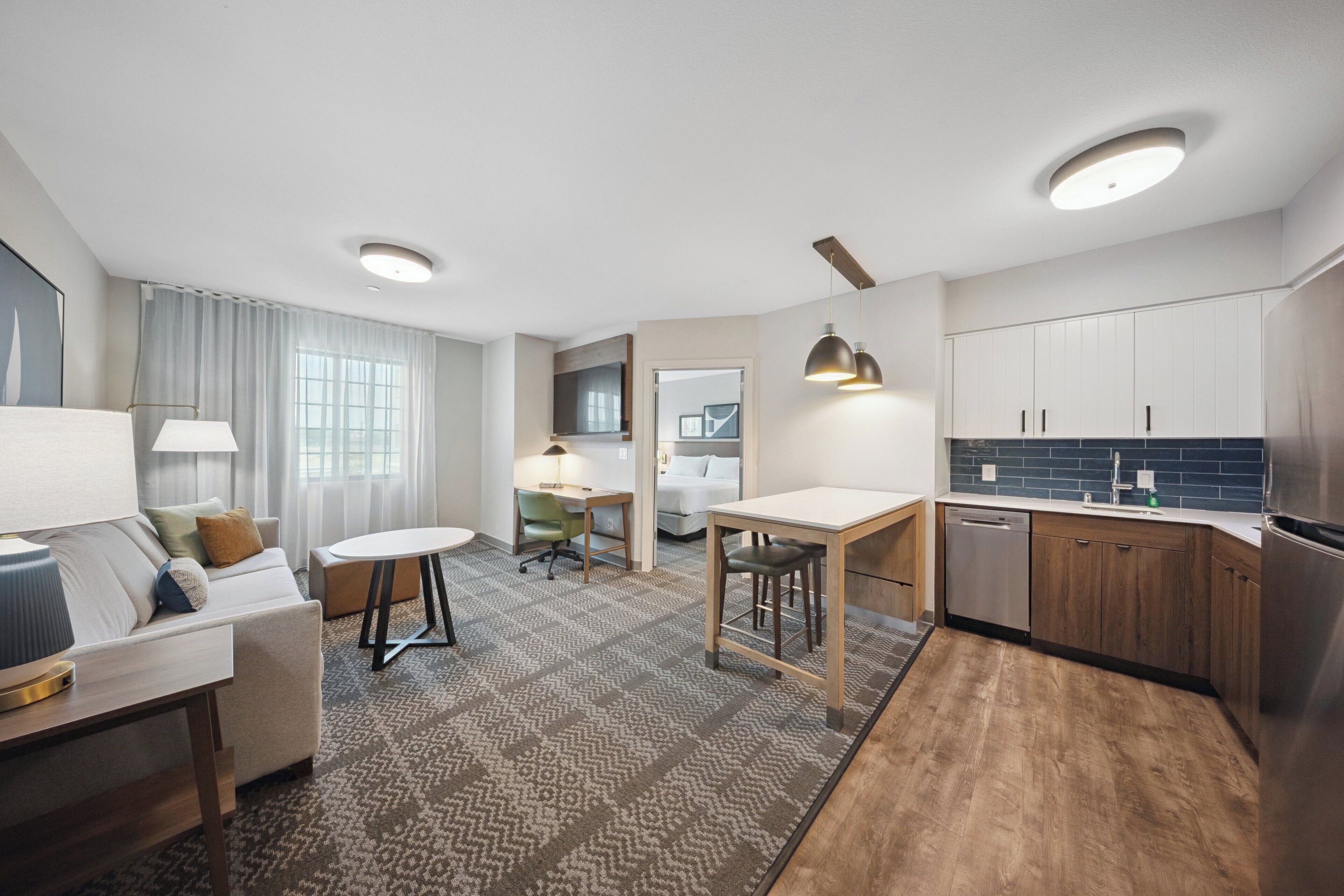 A modern hotel room with a living area, a dining table, a kitchen with wooden cabinets, and a built-in desk.