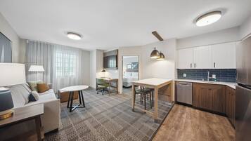 A modern hotel room with a living area, a dining table, a kitchen with wooden cabinets, and a built-in desk.