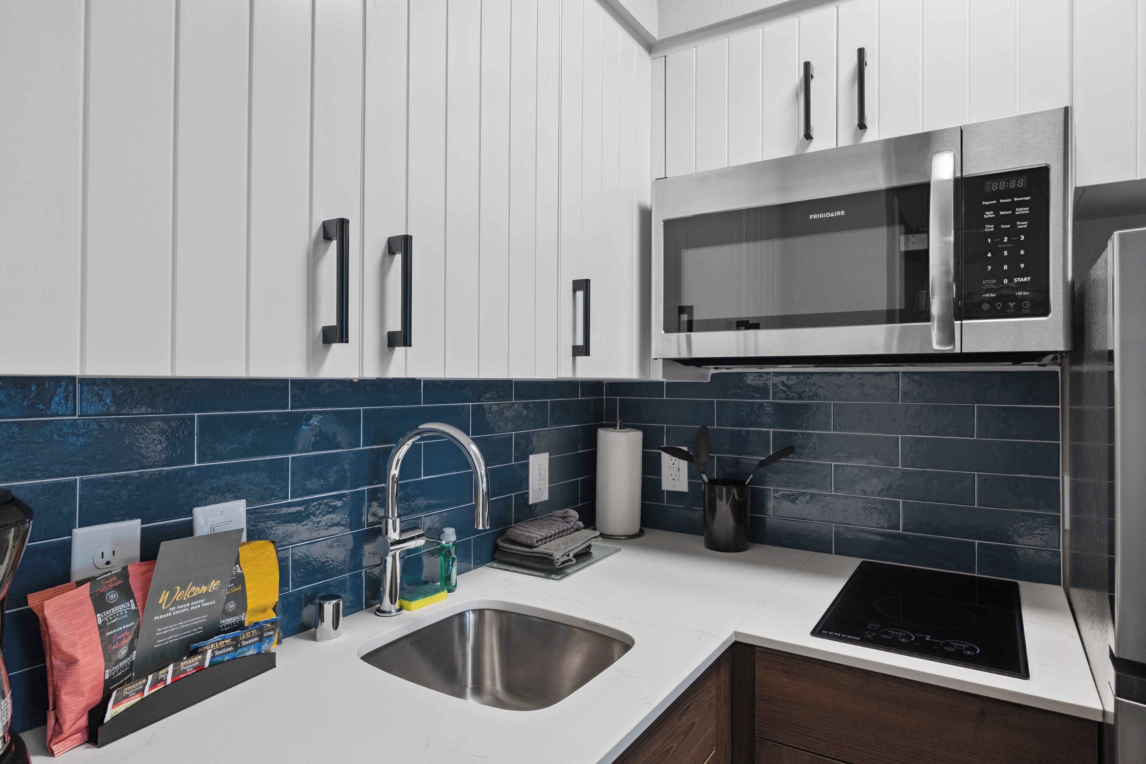 Una cocina moderna con armarios blancos, un lavamanos de acero inoxidable, una placa de inducción negra, un microondas y un salpicadero de azulejos azules.