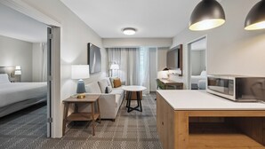 A hotel room with a bed, a sofa, a small dining table, a microwave, and a large mirror. - Staybridge Suites Sacramento - Woodland by IHG (Woodland)