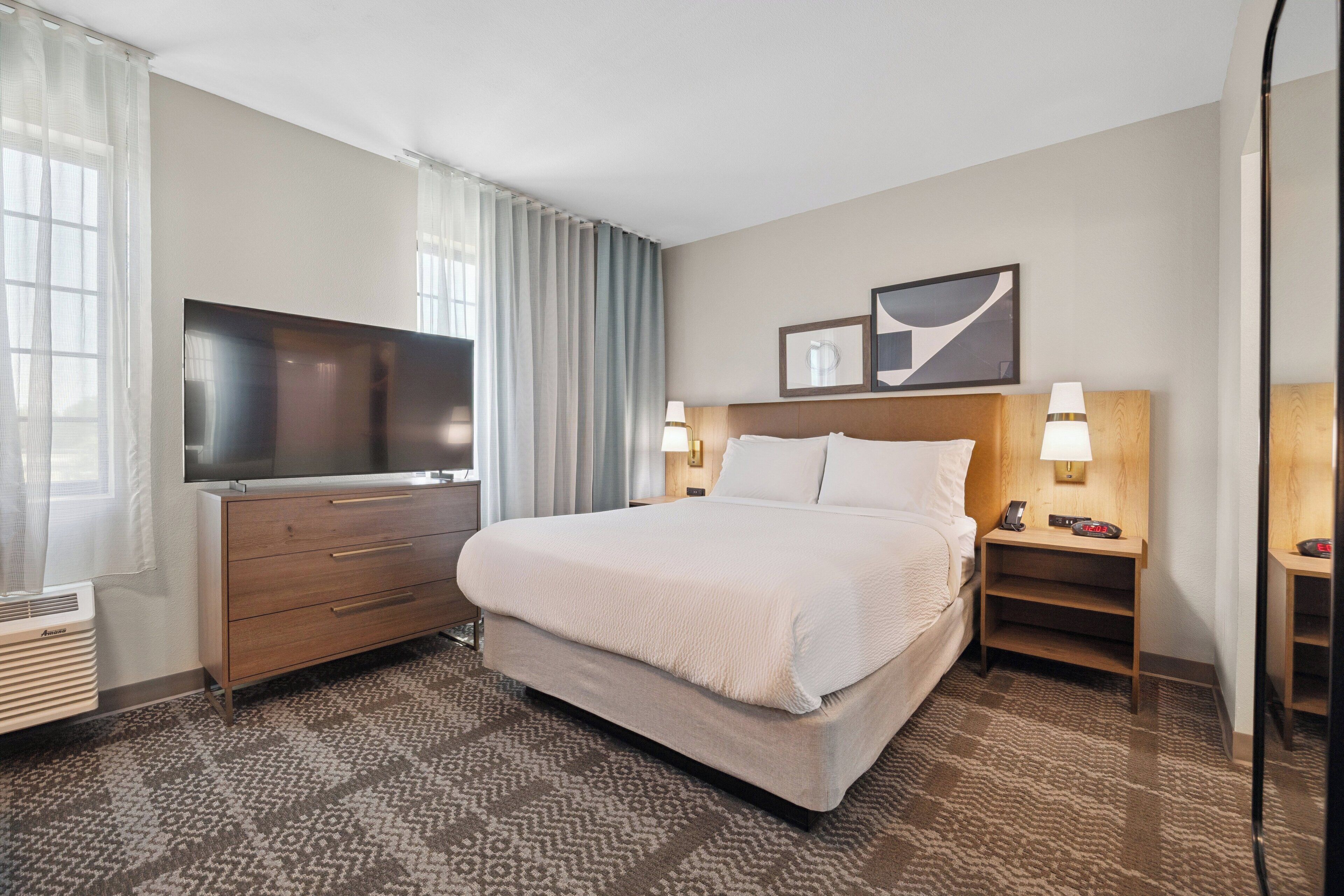 Une chambre d’hôtel avec un grand lit, un téléviseur à écran plat, une commode en bois et une table de chevet avec une lampe.