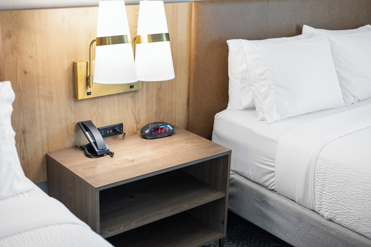 Una habitación de hotel con una mesita de noche de madera, una lámpara de pared, un reloj y una cama con ropa de cama blanca.