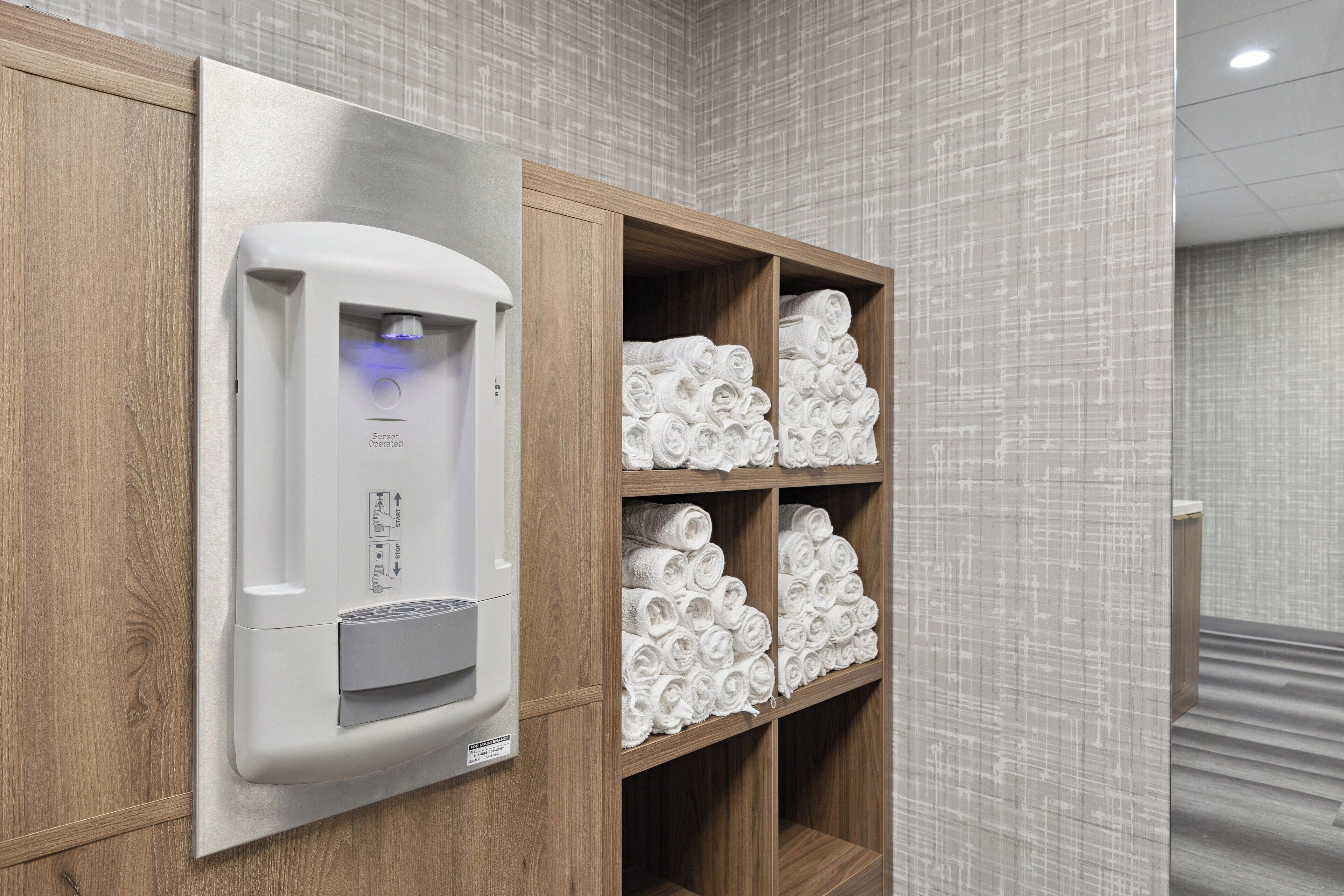 A wall-mounted towel dispenser with a sensor and a wooden shelf with neatly rolled towels.