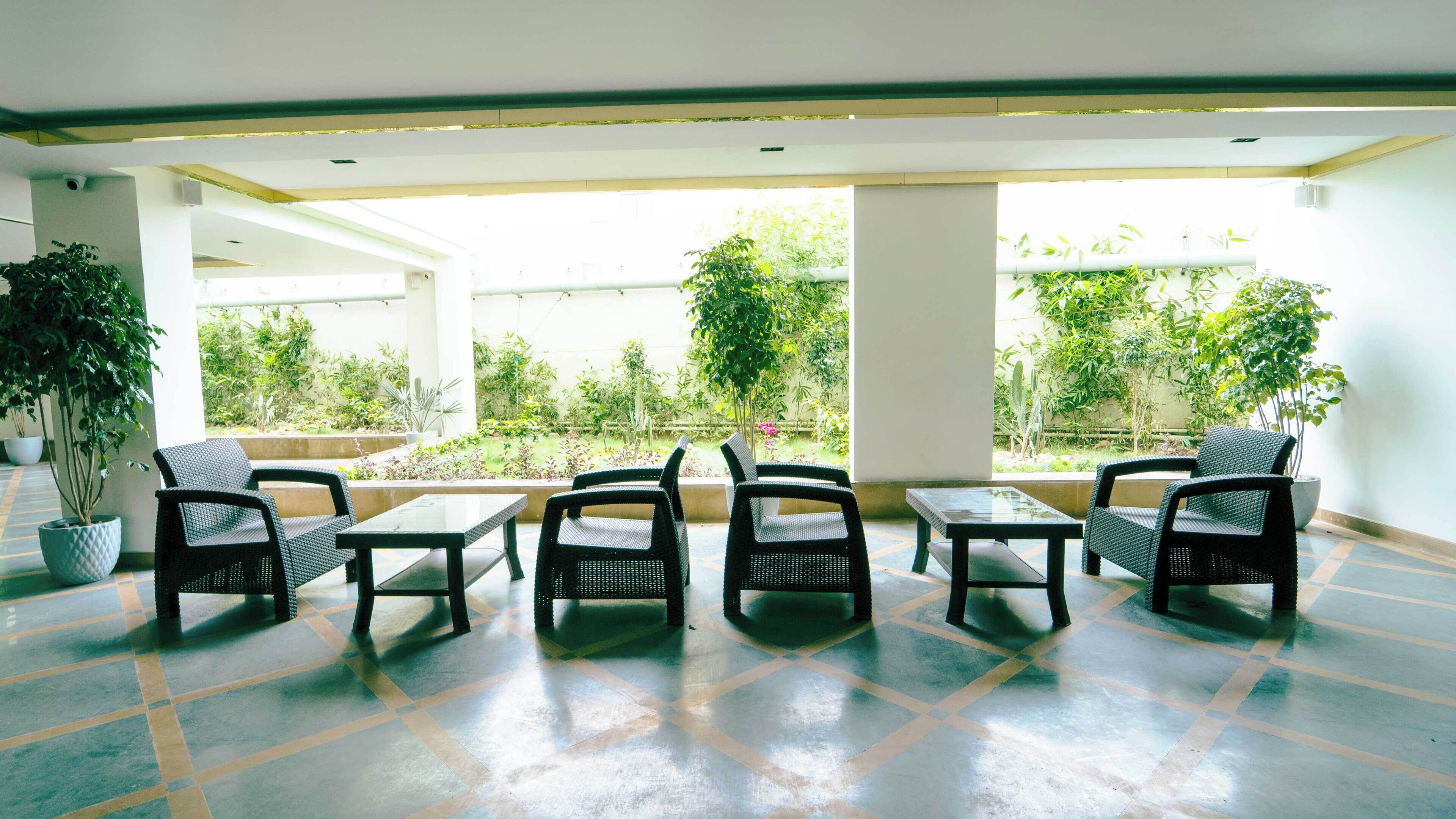 Un vaste espace intérieur doté d’un sol carrelé, de plusieurs fauteuils en osier et de tables disposées pour accueillir les personnes. De grandes fenêtres offrent une vue sur un jardin luxuriant.