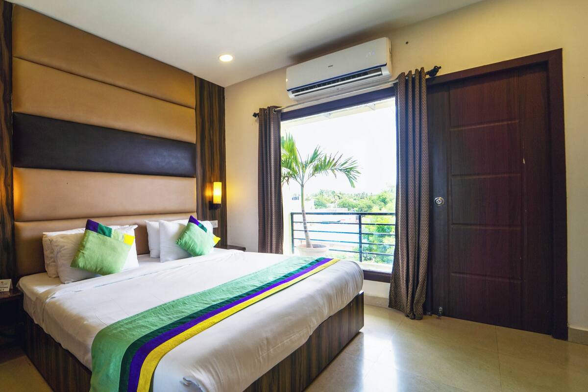 A hotel room with a large bed, a nightstand, a wall-mounted lamp, an air conditioning unit, and a view of the outdoors through a window.
