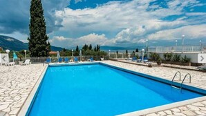 Une piscine rectangulaire bordée de pierres, entourée de chaises longues et de parasols, avec en arrière-plan des montagnes et un ciel nuageux.