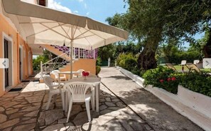 Une terrasse extérieure avec un sol en pierre, des chaises en plastique blanches, une table et un grand parasol.