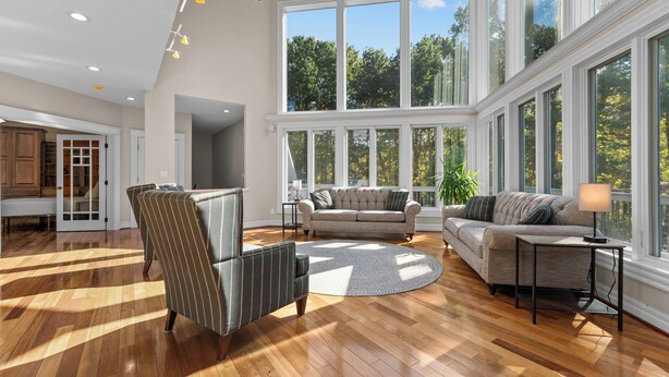 Un salon spacieux doté d’un parquet, d’un fauteuil à rayures, d’un canapé et d’une table basse. De grandes fenêtres offrent une vue sur les arbres et donnent sur un balcon.