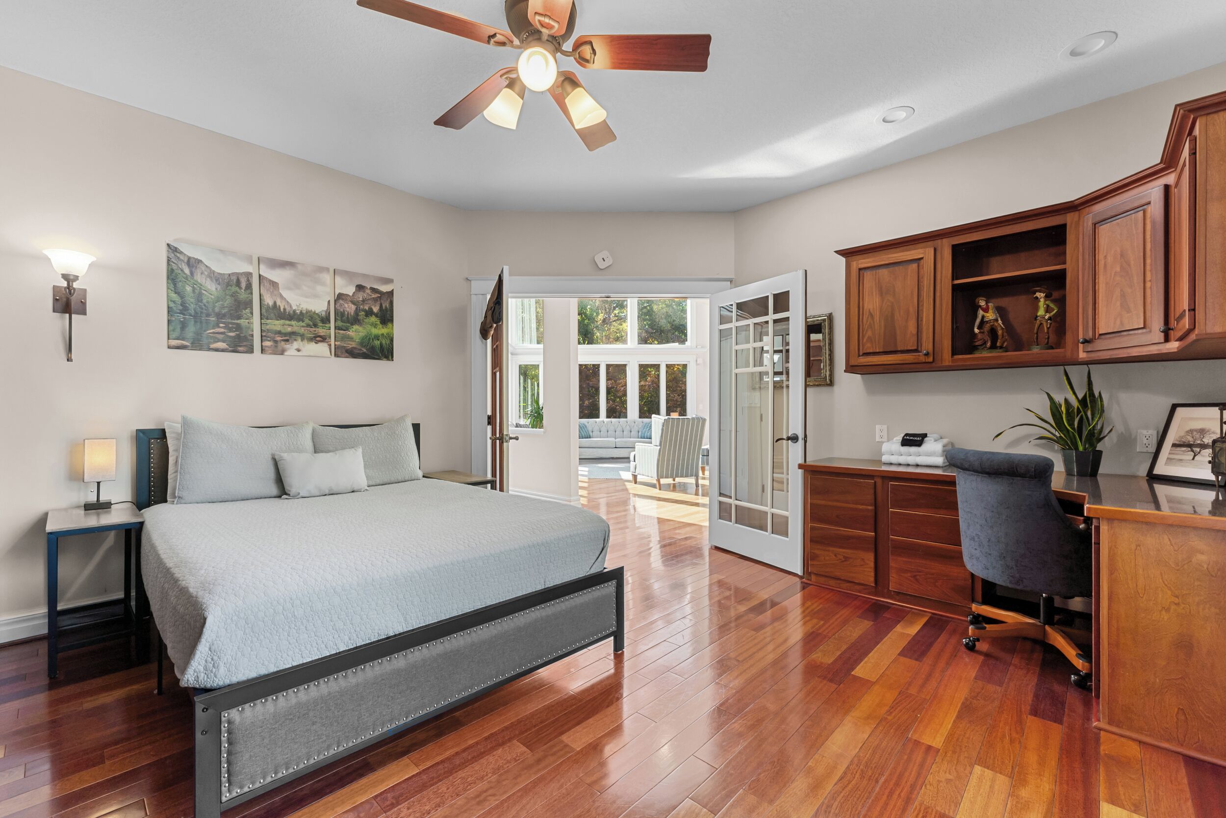 A bedroom with a bed, wooden flooring, a ceiling fan, a desk with a chair, and a view of the outdoors.