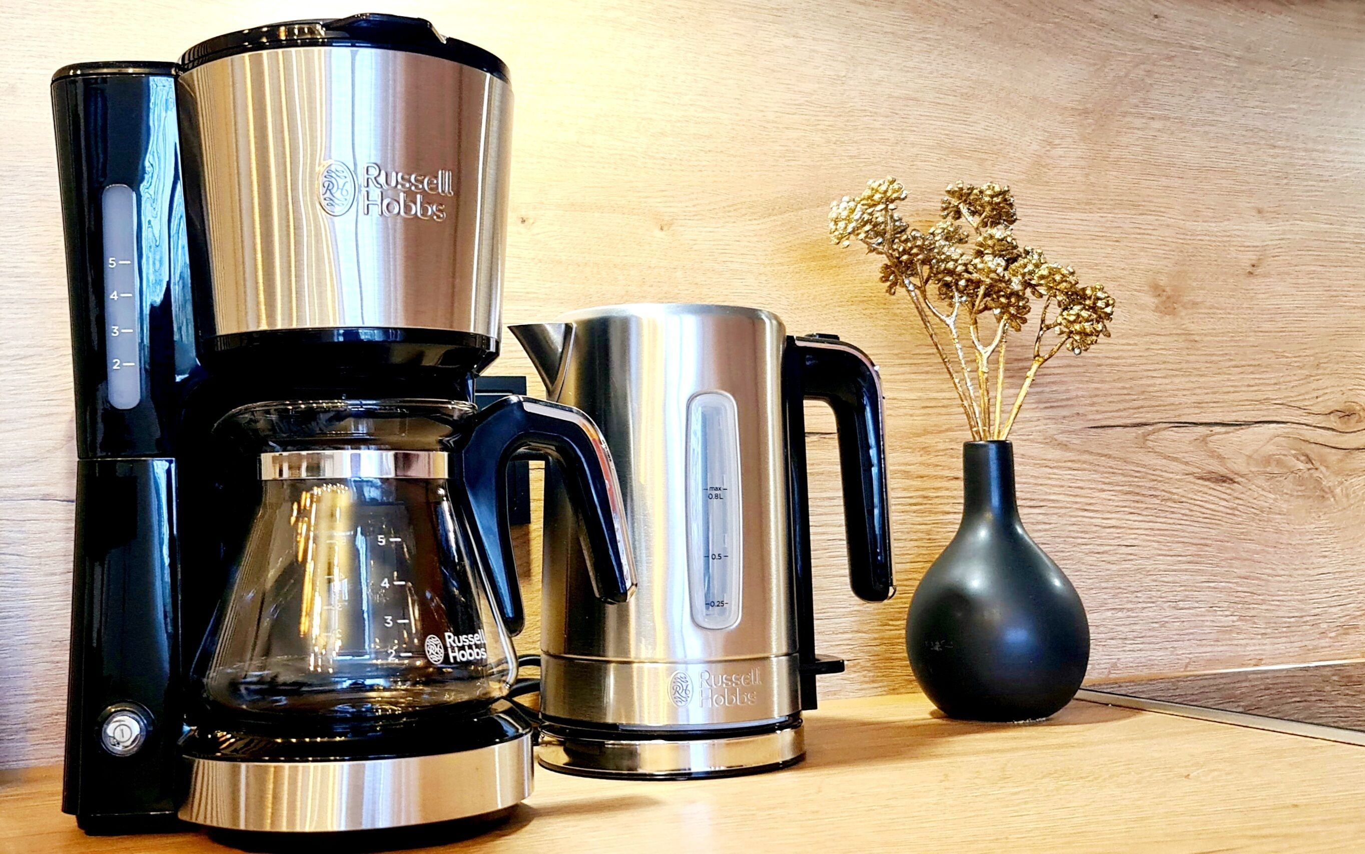A coffee maker, a kettle, and a vase with dried flowers on a wooden surface.
