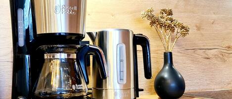 A coffee maker, a kettle, and a vase with dried flowers on a wooden surface.