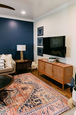 A living room with a dark blue wall, a sofa, a coffee table, a patterned rug, a TV, and a sideboard with a plant. - Lucky Number 7 by Pettigrew Adventures (Memphis)