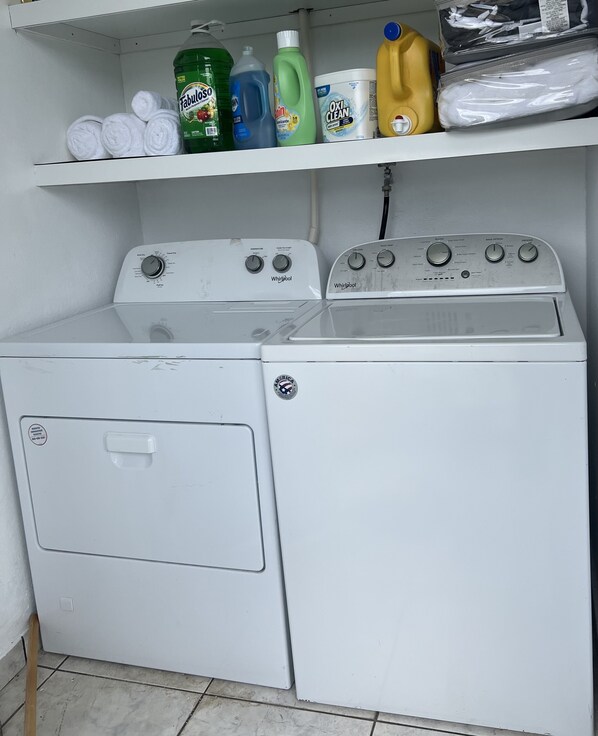 Un área de lavandería con dos lavadoras y secadoras apiladas, un estante arriba con diversos productos de limpieza, y piso de baldosas.