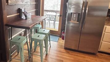 Une cuisine avec une table et des tabourets en bois, un réfrigérateur en acier inoxydable et une porte donnant sur un espace extérieur.
