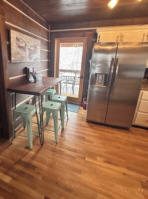 A kitchen with a wooden table and stools, a stainless steel refrigerator, and a door leading to an outdoor area.