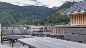 A wooden deck with a sofa and tables, overlooking a forested mountain range.