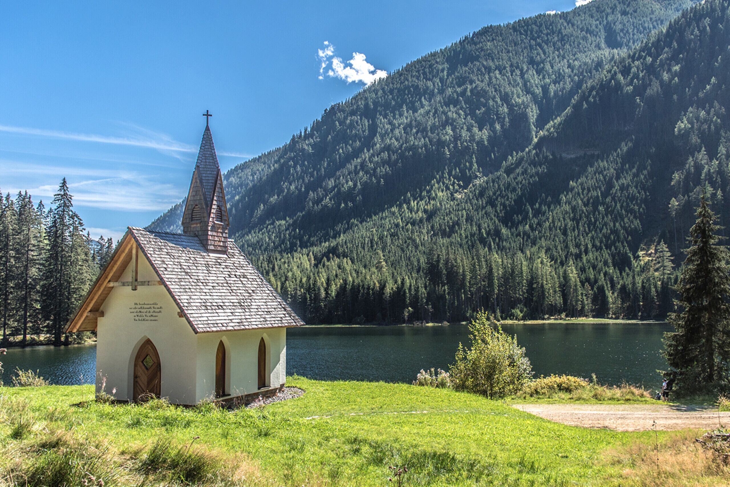 Una iglesia pequeña con un campanario puntiagudo, rodeada por un lago y montañas boscosas.