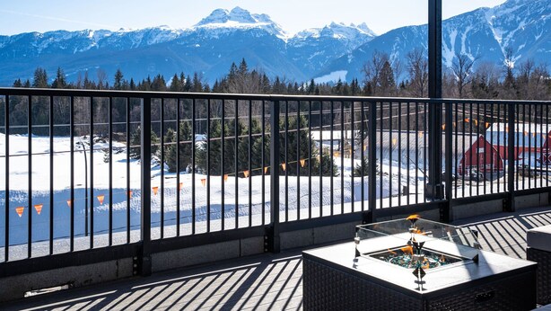 A balcony with a view of snow-covered mountains, a lake, and a red building in the distance. - The Big Deck by Revelstoke Vacations (Revelstoke)