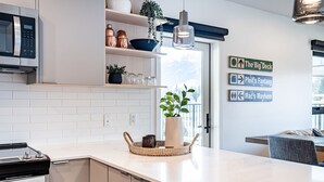 A modern kitchen with white cabinetry, a built-in microwave, and a hanging light fixture. There are decorative items on the shelves and a potted plant on the counter. - The Big Deck by Revelstoke Vacations (Revelstoke)