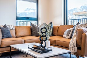 A modern living room with a brown leather sofa, a coffee table with books and a decorative piece, and a view of mountains through large windows.