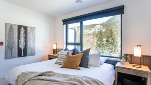 A bedroom with a large bed, a window with a mountain view, and a nightstand with a lamp. - The Big Deck by Revelstoke Vacations (Revelstoke)