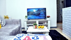 Un salon avec un téléviseur à écran plat posé sur un meuble blanc, un canapé gris, une petite table blanche ornée d’une plante et de fruits, et un tapis noir.
