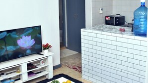 Un salon avec une télévision, une petite table blanche ornée d’une plante et de fruits, un tapis noir et un coin cuisine équipé d’un distributeur d’eau et d’un grille-pain.