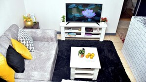 Un salon avec un canapé gris, des coussins noirs et jaunes, une table basse blanche sur laquelle se trouvent des fruits, un téléviseur à écran plat et une petite plante.