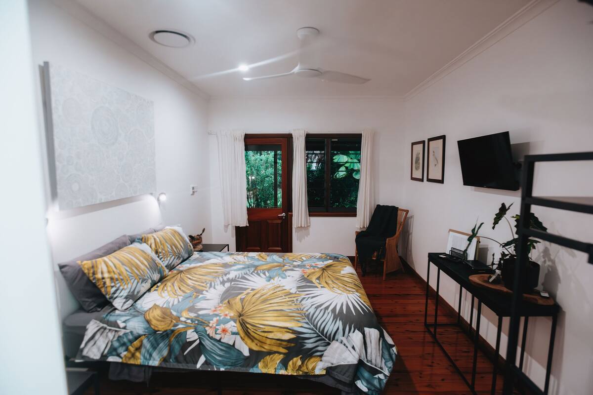 A bedroom with a bed, a desk, a chair, a TV, and a window with curtains.